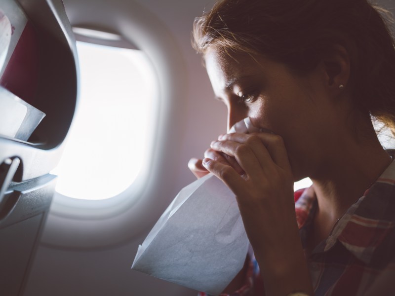 Frau mit Reiseübelkeit sitzt im Flugzeug, sie hält eine Papiertüte vor dem Gesicht