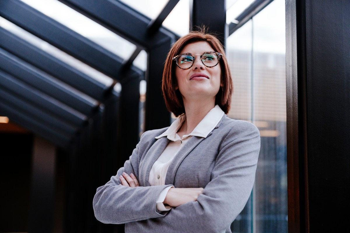 Eine Business-Frau steht mit verschrÃ¤nkten Armen vor einem Fenster.