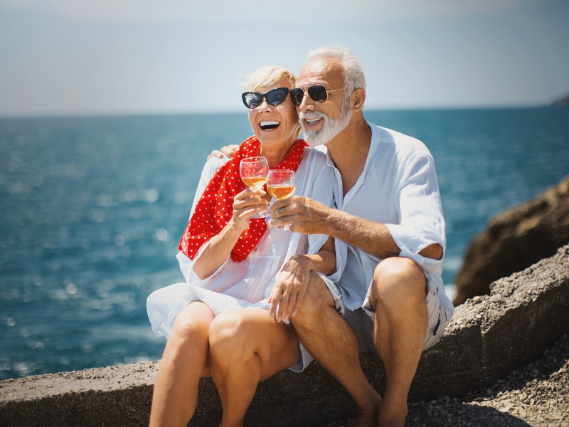 Ein Rentnerpärchen sitzt am Meer und trinkt Wein.