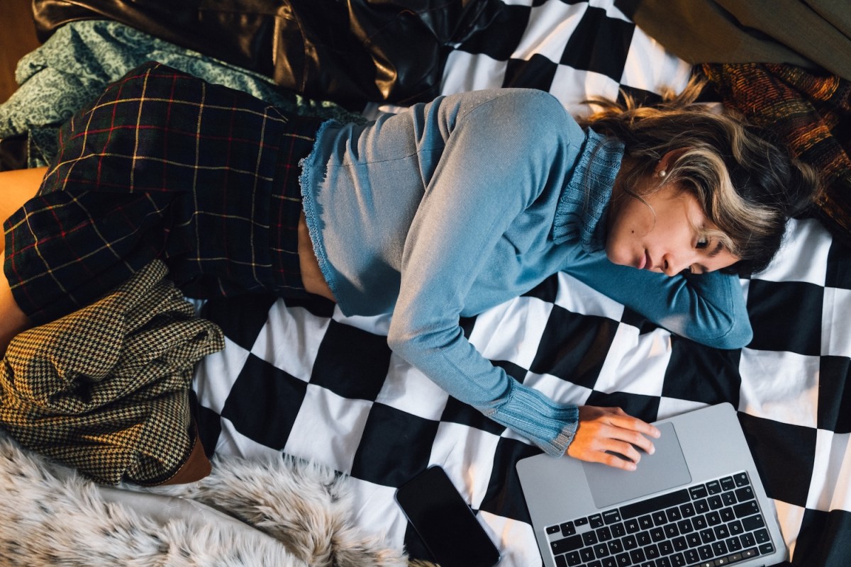 Eine junge Frau liegt auf dem Bett und arbeitet am Laptop.