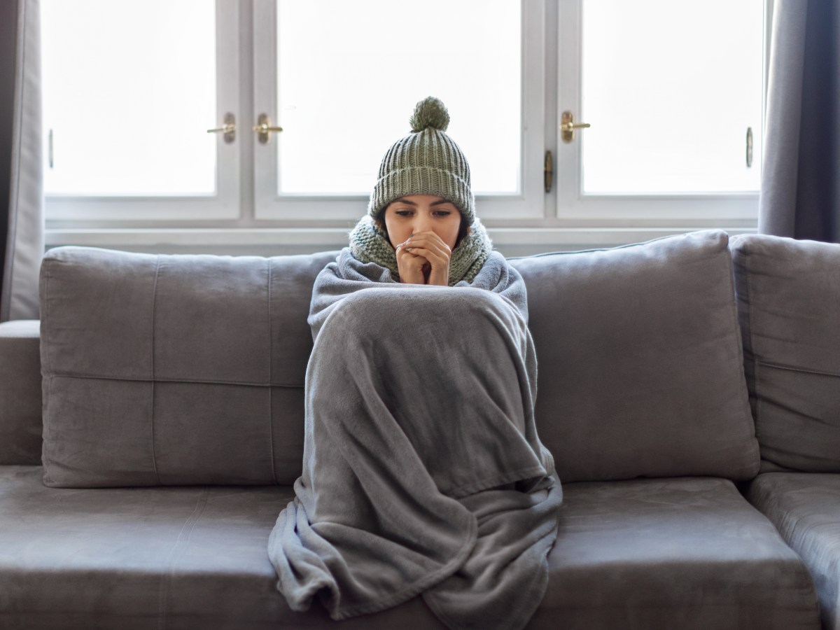 Eine Frau sitzt mit einer decke auf dem Sofa. Sie trÃ¤gt eine MÃ¼nze und friert.