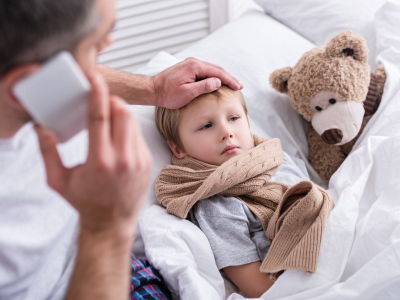 Ein Kind liegt krank im Bett. Der Vater hält die Hand auf seine Stirn.