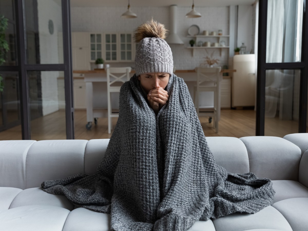 Eine Frau sitzt frierend in ihrer Wohnung. Sie trÃ¤gt eine MÃ¼tze und ich in eine Decke eingewickelt.