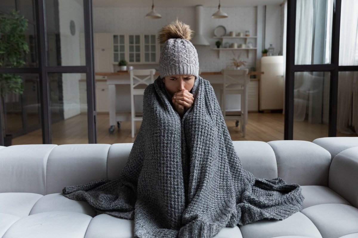 Eine Frau sitzt frierend in ihrer Wohnung. Sie trÃ¤gt eine MÃ¼tze und ich in eine Decke eingewickelt.