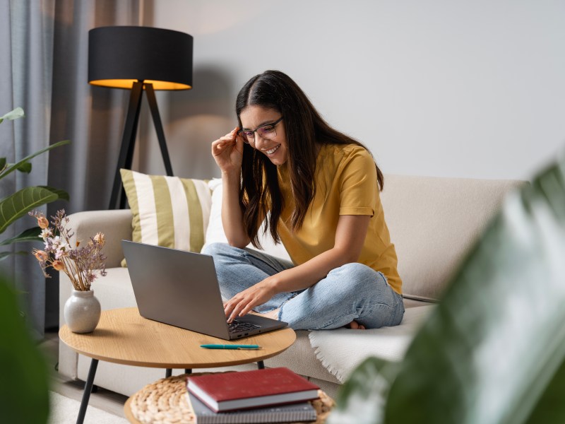 Eine Frau sitzt auf der Couch in ihrem Wohnzimmer und arbeitet am Laptop.