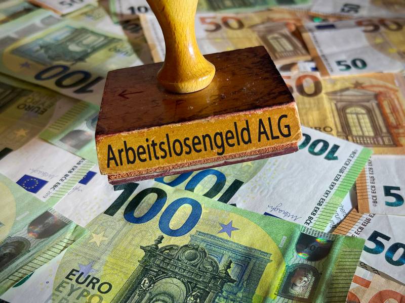 Auf einem Stempel steht das Wort "Arbeitslosengeld ALG". Darunter liegen diverse Geldscheine.