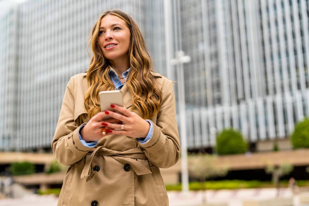 Eine Frau mit Trenchcoat hält ihr Handy in der Hand.