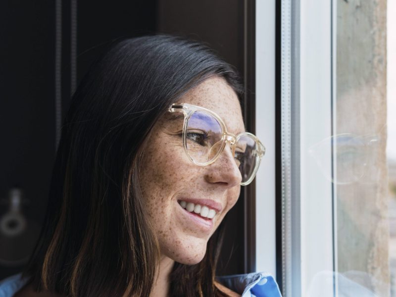 Eine Frau mit Sommersprossen und Brille blickt aus dem Fenster.