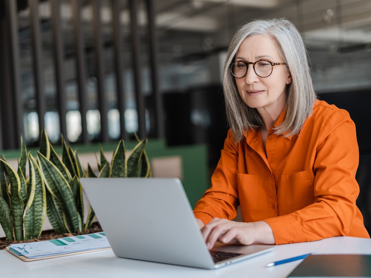 Eine Ã¤ltere Dame mit grauen Haaren sitzt im BÃ¼ro vor einem Laptop.