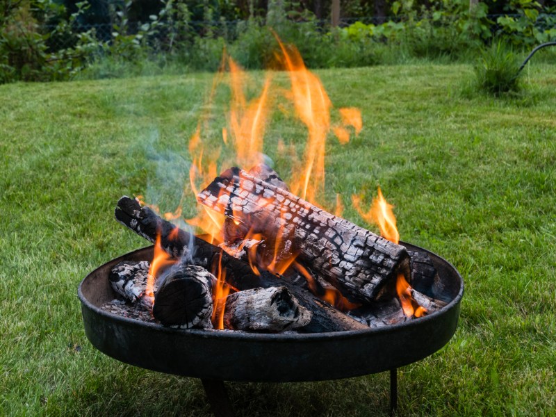 Eine Feuerschale mit brennendem Holz steht in einem Garten.