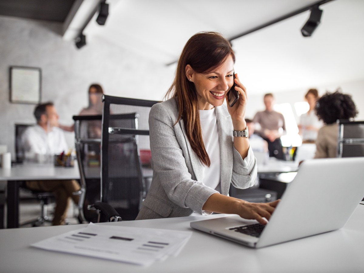 Eine Frau sitzt in einem Großraumbüro und telefoniert. Sie tippt gleichzeitig am Laptop.