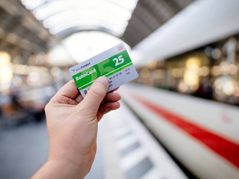 Eine Person hält am Gleis eine BahnCard 25 in der Hand. Im Hintergrund ist ein ICE zu sehen.