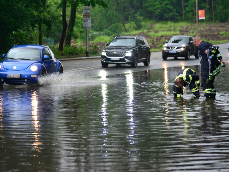 Autos fahren über eine überflutete Straße. Daneben stehen zwei Feuerwehrmänner.