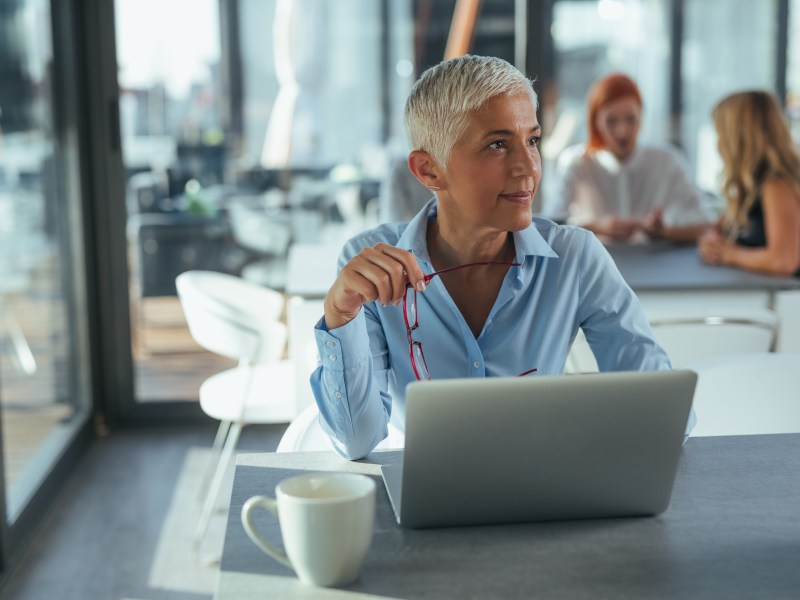 Eine Ã¤ltere Dame mit grauen Haare sitzt im BÃ¼ro vor ihrem Laptop.