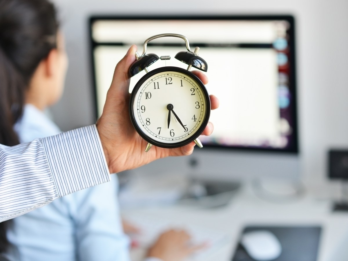 Eine Person hält eine Uhr in der Hand. Dahinter sitzt eine Frau am Computer.