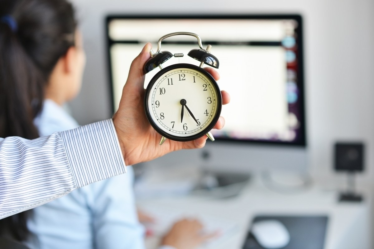 Eine Person hält eine Uhr in der Hand. Dahinter sitzt eine Frau am Computer.