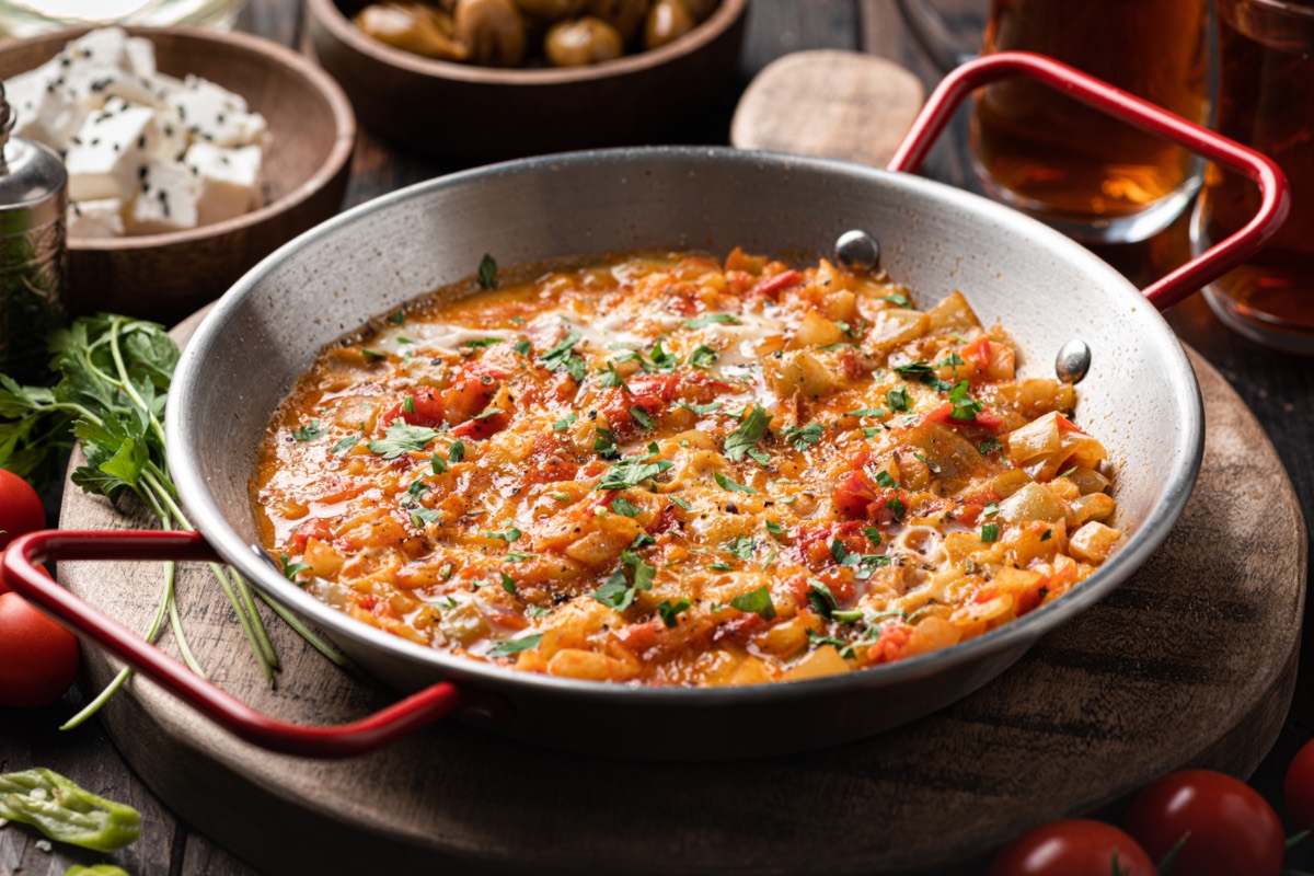 TÃ¼rkisches RÃ¼hrei Menemen in einer Pfanne. Dahinter stehen Schalen mit Zutaten und Flaschen mit Ã–l.