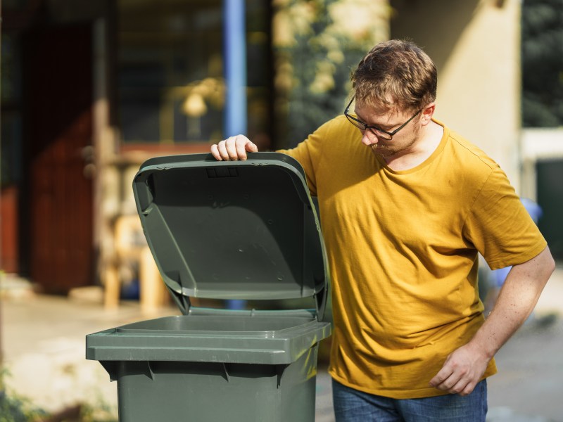 Eine Mann schaut in eine grÃ¼ne MÃ¼lltonne.