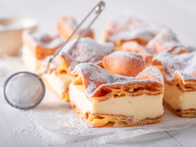 Blätterteigkuchen mit Vanillepudding auf einer weißen Marmorplatte. Darauf liegt ein Sieb, mit dem man den Kuchen mit Puderzucker bestreuen kann.