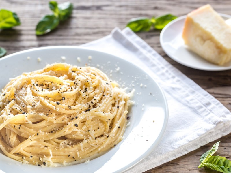 Pasta Cacio e Pepe auf einem weißen Teller, daneben ein Stück Pecorino.