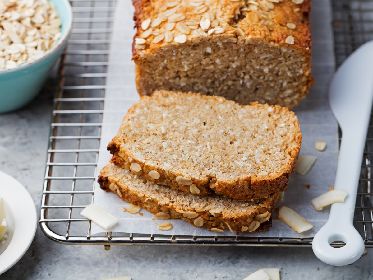 angeschnittenes Haferflocken-Quark-Brot auf einem Brett, daneben eine SchÃ¼ssel Haferflocken und ein Messer.