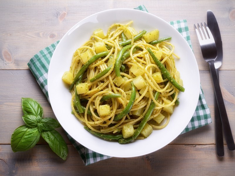 Ligurische Pasta mit Bohnen und Kartoffeln auf einem weiÃŸen Teller. Daneben liegt Besteck und etwas Basilikum.