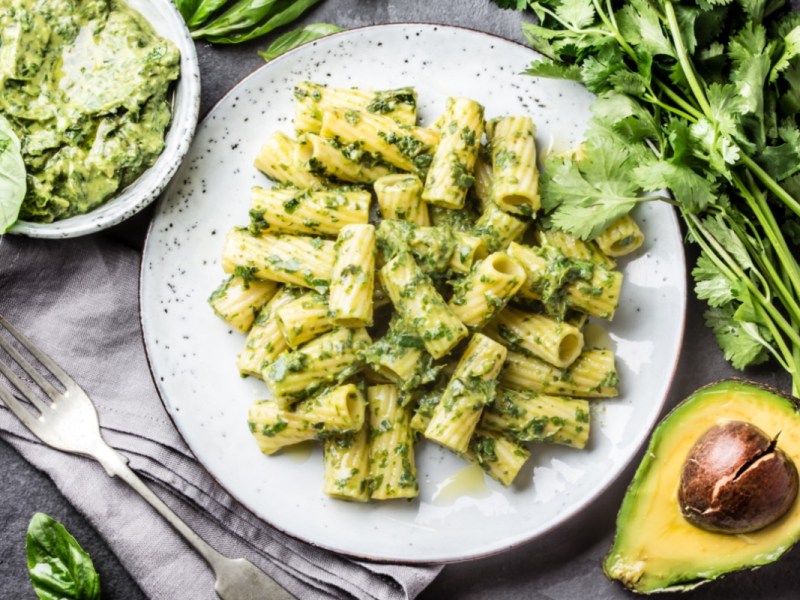 Ein Teller Nudeln mit Avocado-Pesto in der Draufsicht. Daneben eine SchÃ¼ssel Pesto und frische Zutaten.