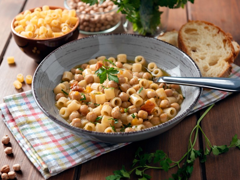 Pasta e Ceci in einer grauen Schüssel. Daneben Schalen mit Nudeln und Kichererbsen sowie Petersilie und zwei Scheiben Brot.