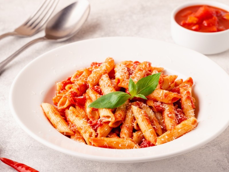 Ein Teller Penne allâ€™arrabbiata. Daneben steht ein SchÃ¤lchen mit Tomaten und es liegt Besteck links vom Teller.