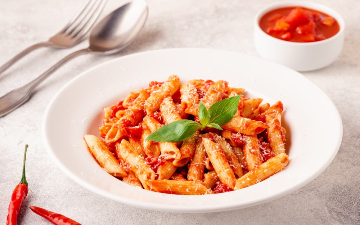 Ein Teller Penne allâ€™arrabbiata. Daneben steht ein SchÃ¤lchen mit Tomaten und es liegt Besteck links vom Teller.