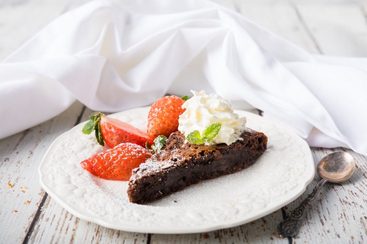 Ein StÃ¼ck schwedischer Schokoladenkuchen Kladdkaka auf einem Teller, garniert mit Erdbeeren und Sahne, daneben ein LÃ¶ffel.