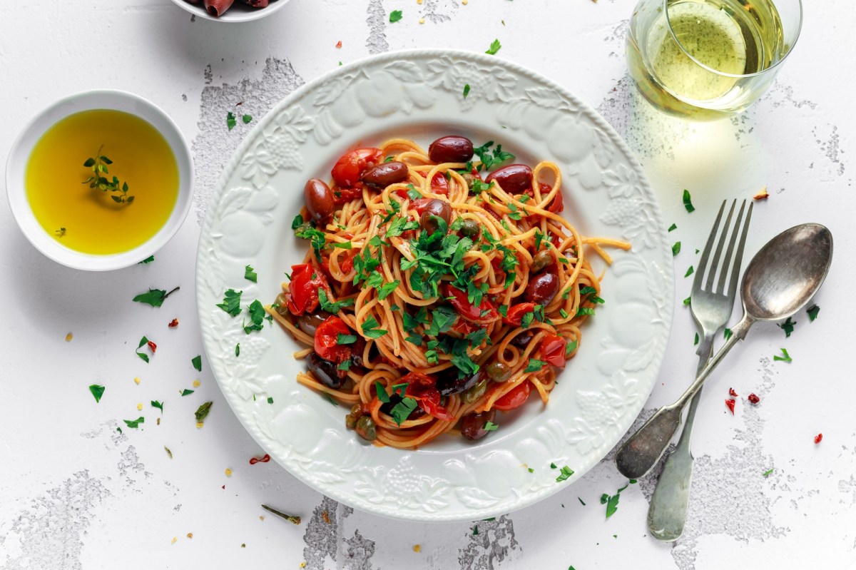 Spaghetti alla Puttanesca auf einem weiÃŸen, gemusterten Teller. Garniert sind sie mit Petersilie und daneben liegen Gabel und LÃ¶ffel sowie OlivenÃ¶l und ein Glas WeiÃŸwein.