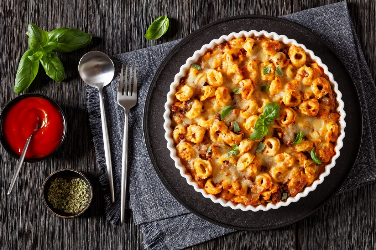 Tortellini-Auflauf in einer geriffelten Auflaufform auf einem schwarzen Tablett. Darunter ein graues Platzdeckchen, LÃ¶ffel und Gabel und TomatensoÃŸe. Draufsicht.