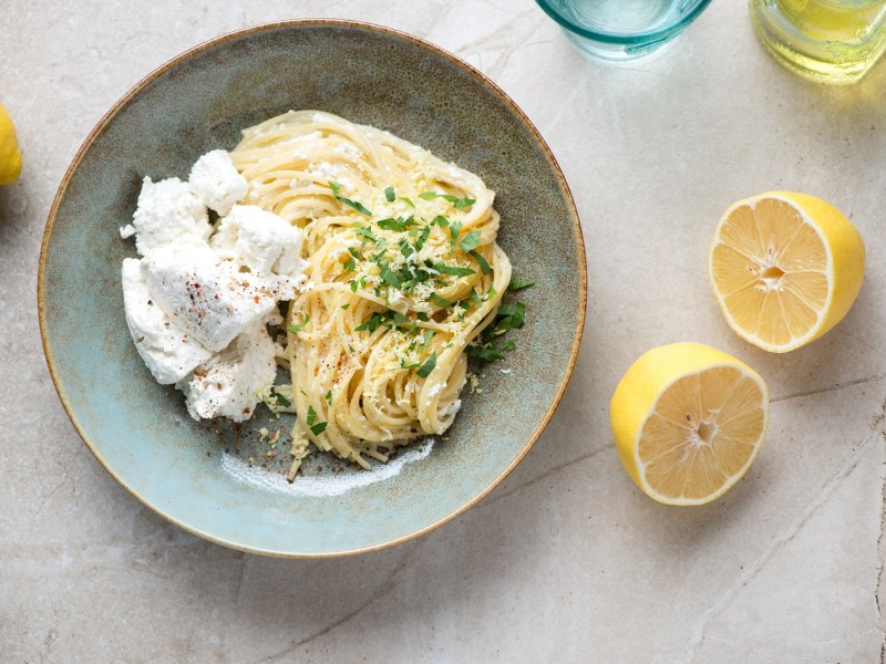 Eine Portion Zitronen-Ricotta-Pasta auf einem blauen Teller. Daneben liegt eine aufgeschnittene Zitrone.