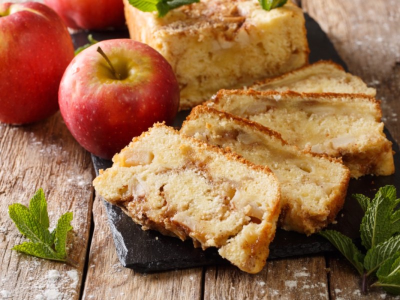 Mehrere StÃ¼cke Apfelbrot, die gefÃ¤chert vor dem restlichen Kuchen liegen. Daneben zwei Ã„pfel.