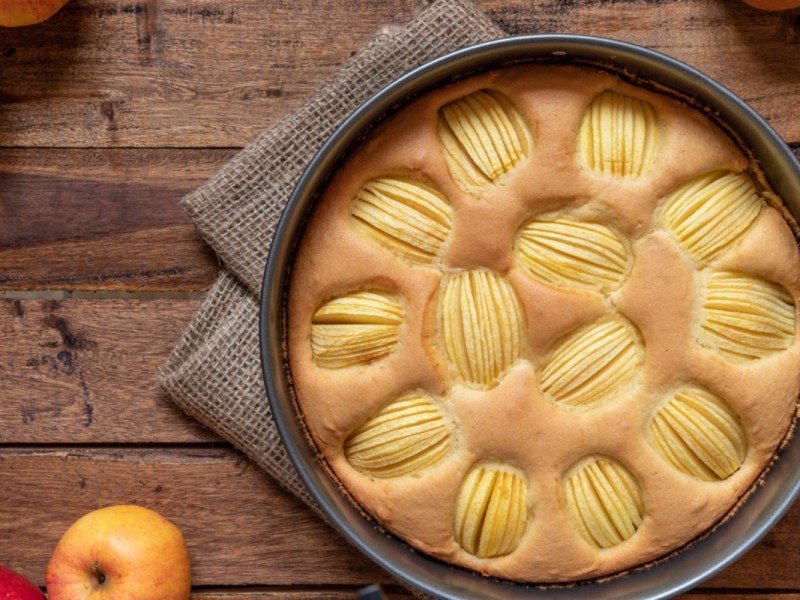 Ein Apfelkuchen ohne Zucker in der Backform von oben fotografiert. Es liegen frische Ã„pfel daneben.