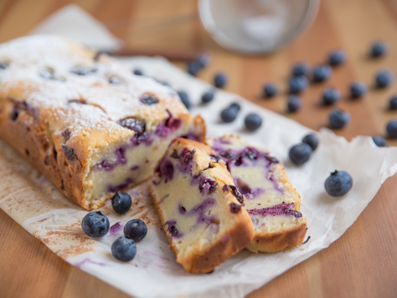 Blaubeer-Joghurt-Kuchen auf Backpapier mit zwei abgeschnittenen StÃ¼ckchen.