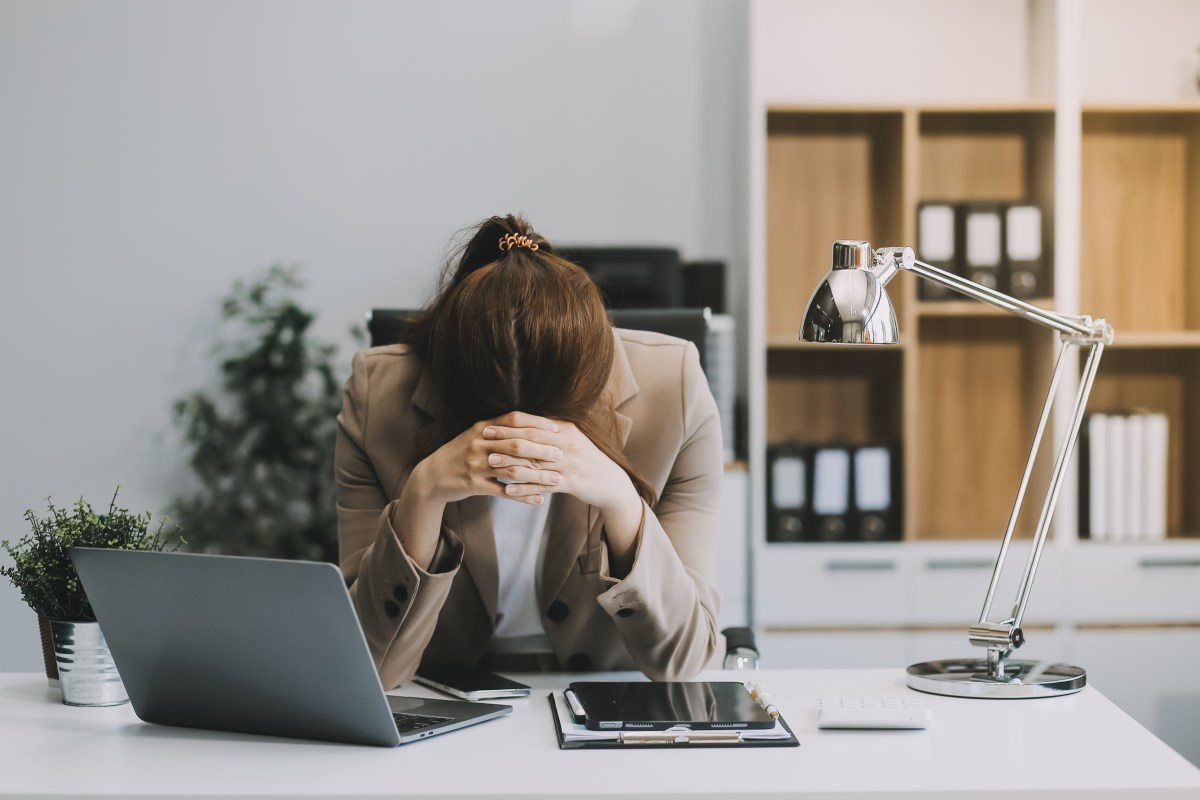 Eine junge Frau sitzt im Büro und ist erschöpft. Sie legt den Kopf auf ihre Hände.