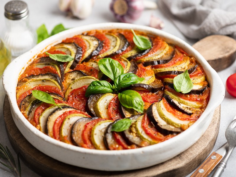 franzÃ¶sischer GemÃ¼seauflauf Tian de LÃ©gumes in einer runden Auflaufform auf einem Holzbrett, drumherum viele Zutaten.