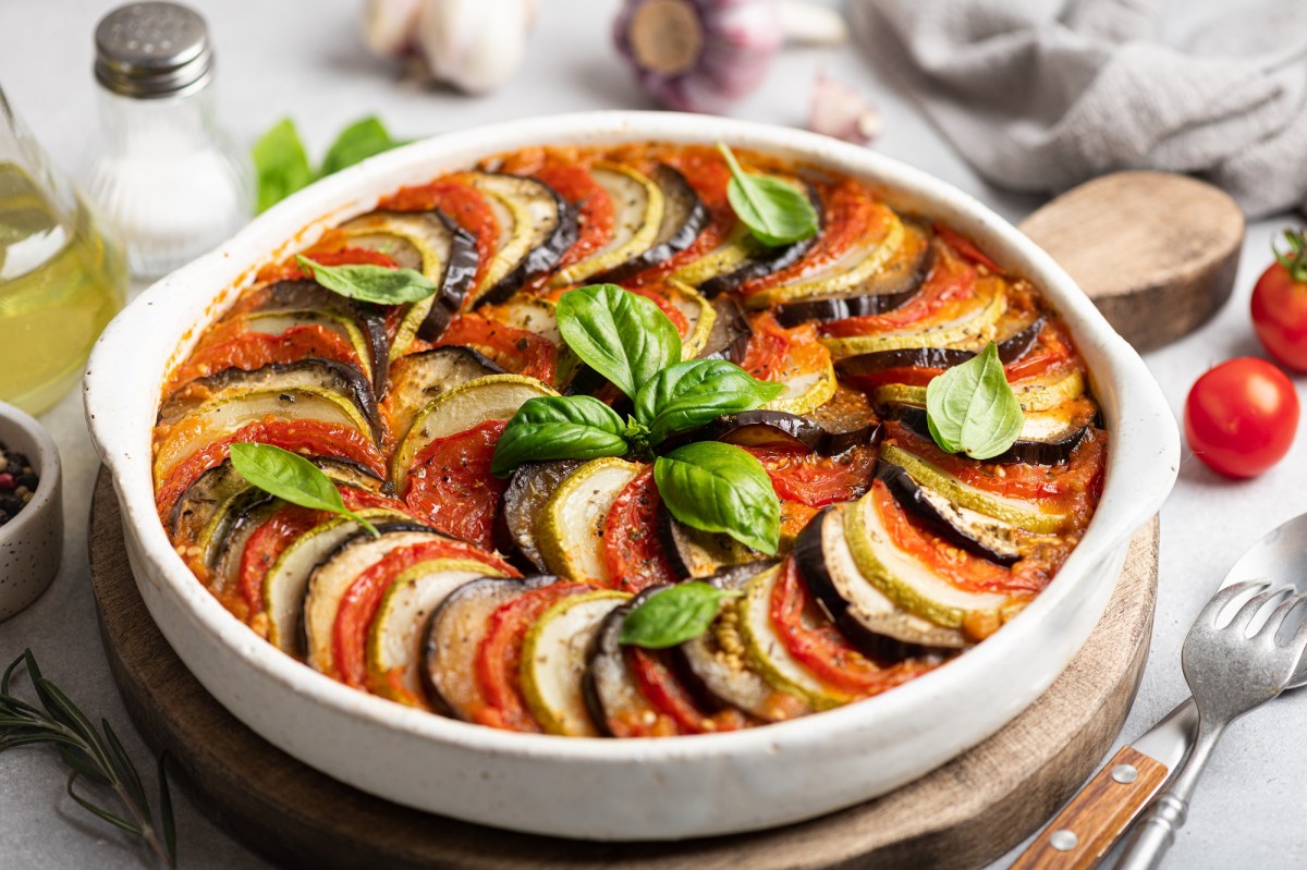 franzÃ¶sischer GemÃ¼seauflauf Tian de LÃ©gumes in einer runden Auflaufform auf einem Holzbrett, drumherum viele Zutaten.