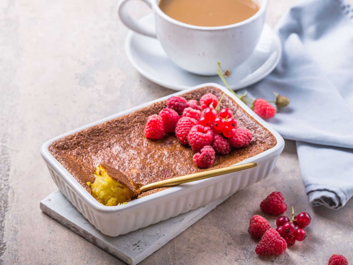 Gebackener Milchreis in einer Auflaufform, angestochen mit einem LÃ¶ffel. Einige Himbeeren liegen als Dekoration auf dem Auflauf und daneben. Im Hintergrund steht eine Kaffeetasse.