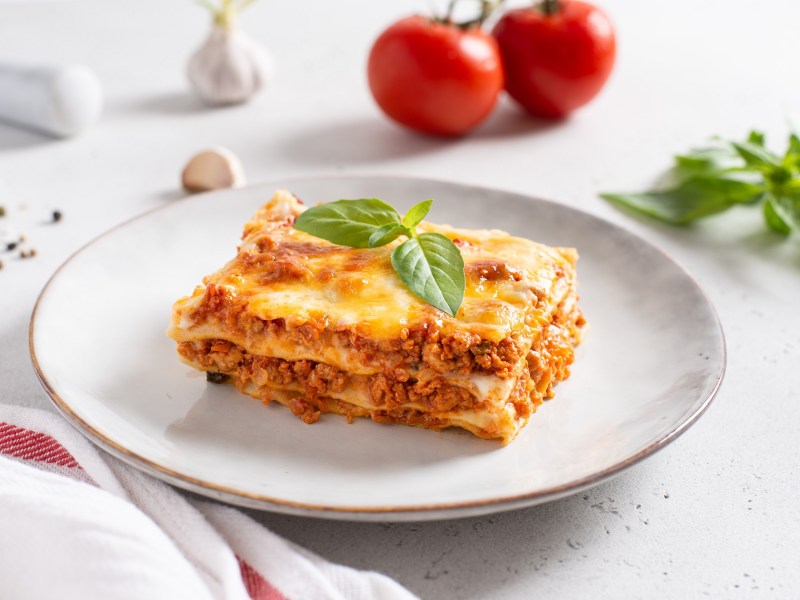 Ein StÃ¼ck Lasagne mit Schmand auf einem Teller, garniert mit Basilikum, dahinter 2 Tomaten.