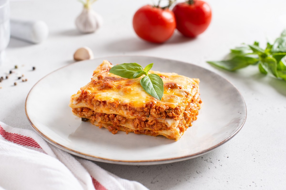 Ein StÃ¼ck Lasagne mit Schmand auf einem Teller, garniert mit Basilikum, dahinter 2 Tomaten.