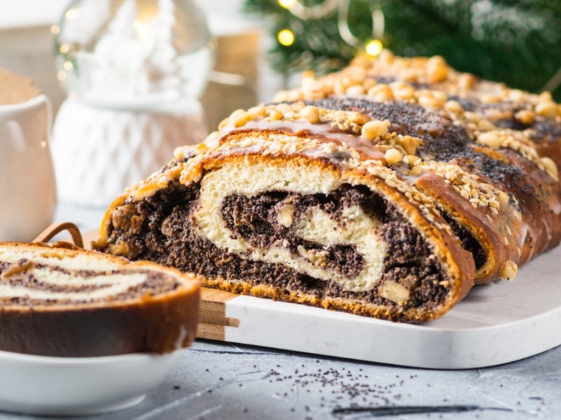 Ein aufgeschnittener Mohnstriezel mit WalnÃ¼ssen und Streuseln, der auf einer lÃ¤nglichen Servierplatte liegt. Links davor liegt ein StÃ¼ck auf einem kleinen Teller. Im Hintergrund sind ein MilchkÃ¤nnchen, eine Schneekugel und TannengrÃ¼n mit einer Lichterkette zu sehen.