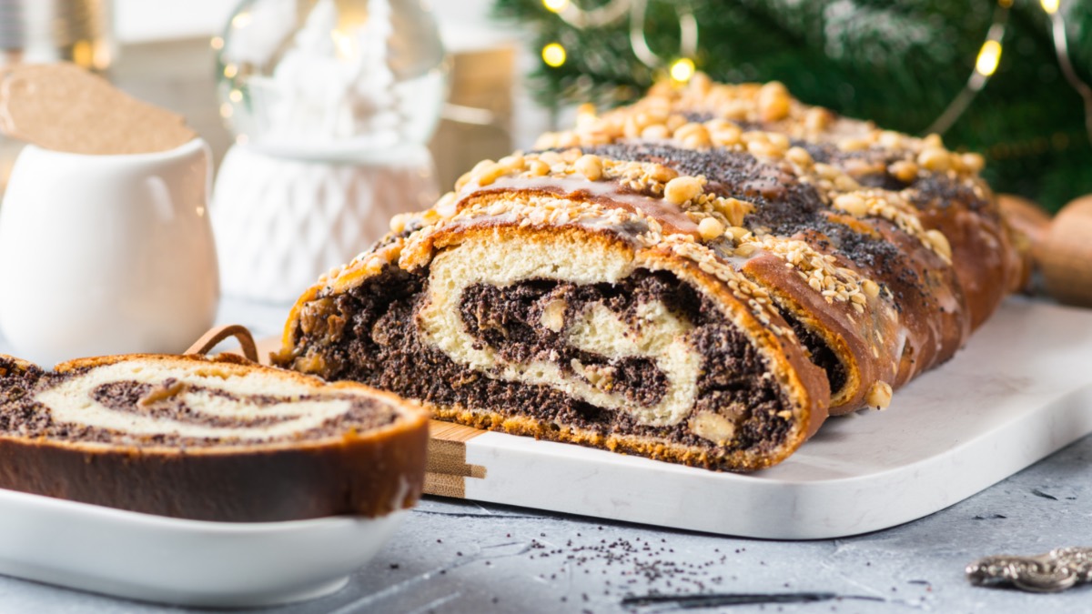 Ein aufgeschnittener Mohnstriezel mit WalnÃ¼ssen und Streuseln, der auf einer lÃ¤nglichen Servierplatte liegt. Links davor liegt ein StÃ¼ck auf einem kleinen Teller. Im Hintergrund sind ein MilchkÃ¤nnchen, eine Schneekugel und TannengrÃ¼n mit einer Lichterkette zu sehen.