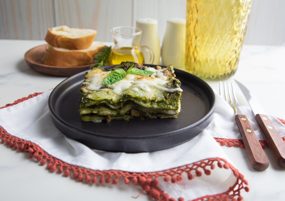 Pesto-Lasagne auf einem schwarzen Teller. Dahinter stehen ein Teller Brot, eine Flasche OlivenÃ¶l und Salz nud Pfeffer.