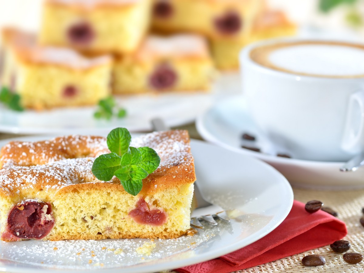 Quark-Kirsch-Kuchen auf einem weiÃŸen Teller. Im Hintergrund steht mehr Kuchen, daneben eine Tasse Cappuccino.