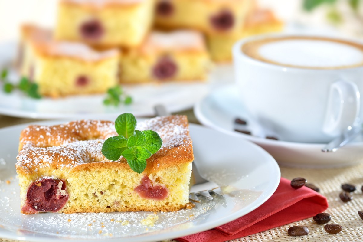 Quark-Kirsch-Kuchen auf einem weiÃŸen Teller. Im Hintergrund steht mehr Kuchen, daneben eine Tasse Cappuccino.