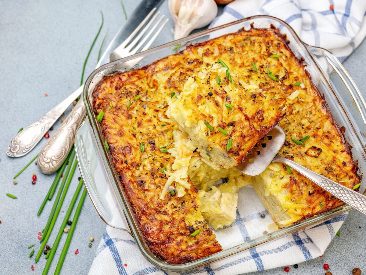 RÃ¶sti-Schnitzel-Auflauf in einer glÃ¤sernen Ofenform. Darunter liegt ein kariertes KÃ¼chentuch und Besteck sowie FrÃ¼hlingszwiebeln als Deko daneben.