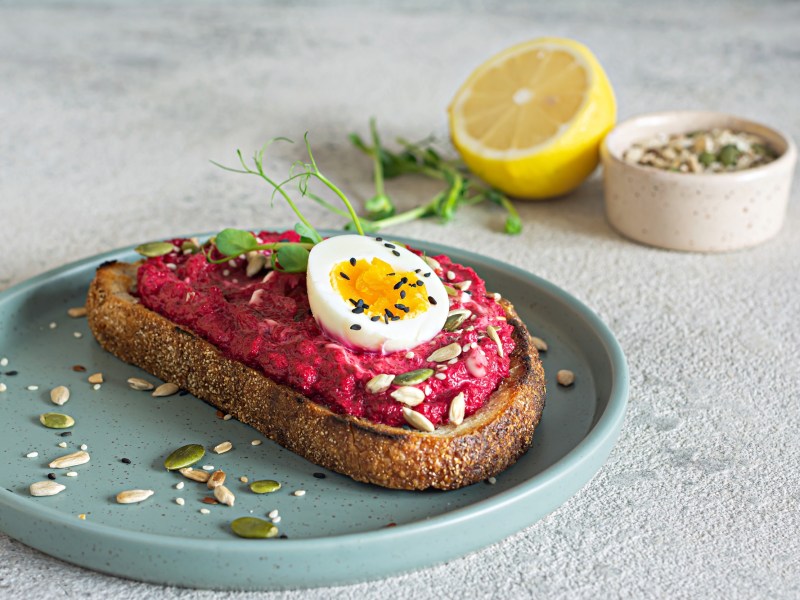 Eine Scheibe Brot mit Rote-Bete-Aufstrich mit FrischkÃ¤se und Ei auf einem Teller, garniert mit Kernen, im Hintergrund eine halbe Zitrone und eine Schale Kerne.